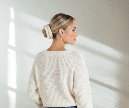 Woman wearing a cream white knit scrunchie and cream white sweater standing against a white wall with sunlight casting shadows.