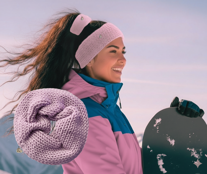 Woman in winter clothing with a pink knit headband and scrunchie holding a snowboard outdoors.