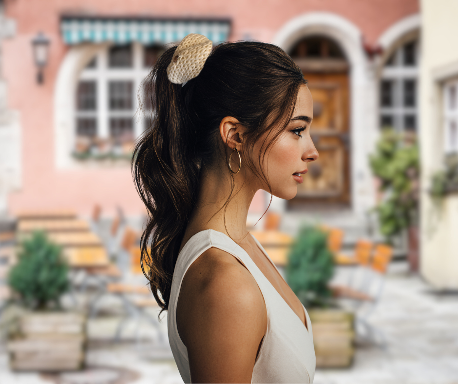 Woman with cream white knit wool scrunchie on her ponytail standing in front of a building with plants and furniture.