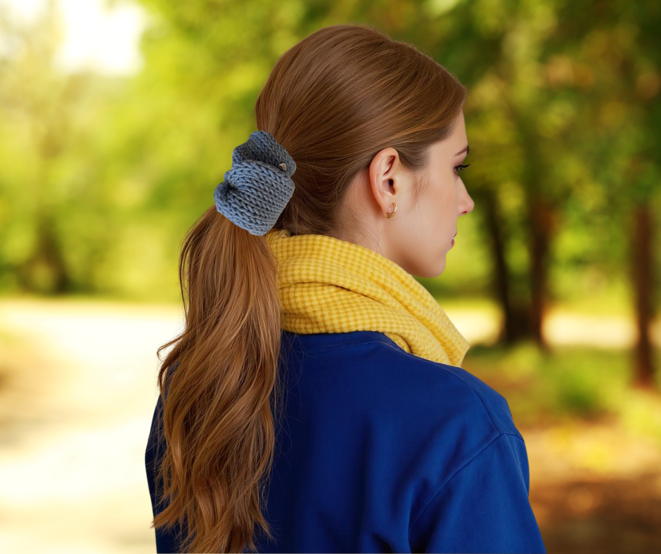 Woman with a blue scrunchie in her hair, wearing a yellow scarf and blue coat, standing in a park.