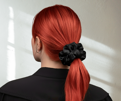 Model with red hair tied in a ponytail with a Onyx black silk scrunchie, wearing a black shirt on light background.