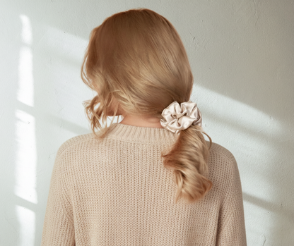 Person with a silk champagne  colored scrunchie in their hair against a white wall